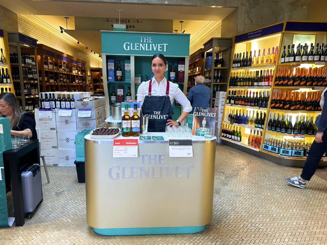 Screenshot 2024-11-26 111708 Brand ambassador at a Glenlivet whisky sampling table, showcasing premium whisky bottles and branded materials in a sleek setup.