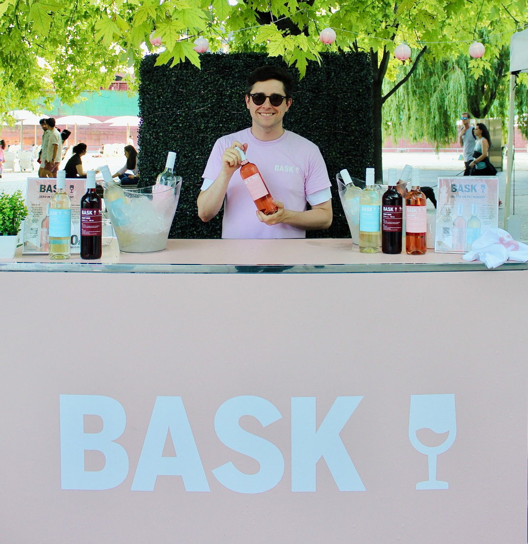 Brand ambassador smiling while holding a Bask 0 Sugar Wine sample at a vibrant beach event, highlighting the refreshing drink.